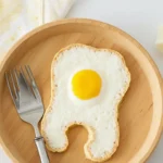 A fun breakfast idea for babies featuring a sunny-side-up egg nestled inside a slice of bread cut into a cute shape, served on a wooden plate with a fork.