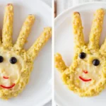 Creative baby finger food arrangement shaped like a hand, made from millet and banana puree with blueberry eyes and a smiley face.