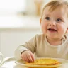 A smiling woman in a modern kitchen using a food processor to prepare fresh baby food with healthy ingredients.