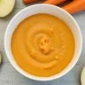 A smooth and nutritious carrot and apple puree in a white bowl, surrounded by fresh carrots and apple slices.